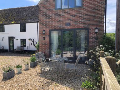 An outdoor seating area with metal chairs and a table in front of a brick building with large windows at Little Stables Cottage in Spetisbury