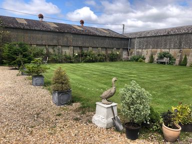 A garden with a lawn, potted plants, a statue of a bird on a pedestal, and a gravel path at Little Stables Cottage in Spetisbury