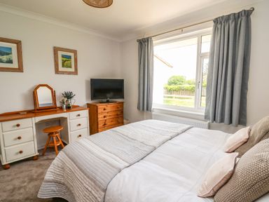 A bedroom with a bed a dressing table a stool a chest of drawers a television and a window with curtains at Bryn Alaw in Newport Pembrokeshire