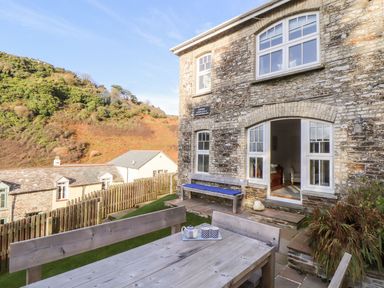 An outdoor patio with wooden table and benches near a stone building with multiple windows and a green hillside at Cliffside in Trebarwith Strand near Tintagel