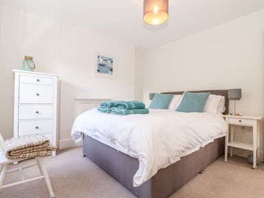 A bedroom with a double bed with blue pillows and folded towels a white dresser and bedside table with lamps at Cliffside in Trebarwith Strand near Tintagel