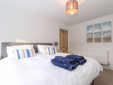 A bedroom with a double bed white bedding blue striped pillows folded blue towels a wooden door and a triptych seascape painting on the wall at Cliffside in Trebarwith Strand near Tintagel