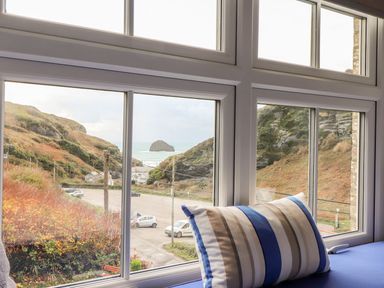 A window seat with a striped pillow overlooking a coastal landscape with hills cars and a rock in the sea at Cliffside in Trebarwith Strand near Tintagel