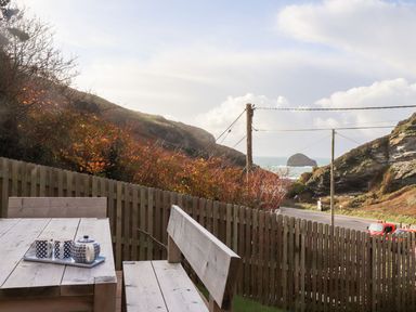 An outdoor wooden table and bench set with a tea set on the table near a wooden fence overlooking a coastal road and rocky hills at Cliffside in Trebarwith Strand near Tintagel