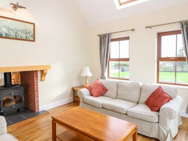 A living room with a beige sofa with red cushions a wooden coffee table a wood burning stove and two windows with curtains at Ballycahane in Castletownshend County Cork