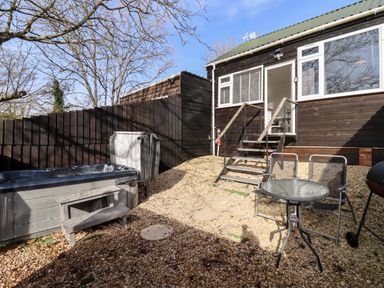 A small outdoor patio area with a hot tub a glass table with two chairs and stairs leading to a wooden house at Stillness in Blandford Forum