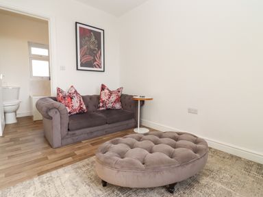 A living room with a gray sofa with floral pillows a round tufted ottoman and a side table next to an open door showing a bathroom at Stillness in Blandford Forum