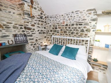 A bedroom with a stone wall a bed with blue pillows a patterned blanket and shelves with kitchen items at Dyffryn Cottage in Aberporth