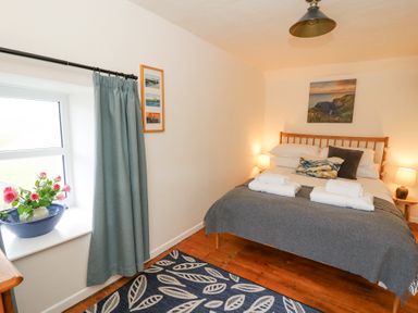 A bedroom with a double bed bedside tables lamps a window with blue curtains a blue patterned rug and flowers in a bowl on the windowsill at Dyffryn Cottage in Aberporth