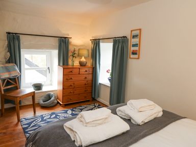 A bedroom with two windows blue curtains a wooden chair a chest of drawers a lamp and towels on the bed at Dyffryn Cottage in Aberporth