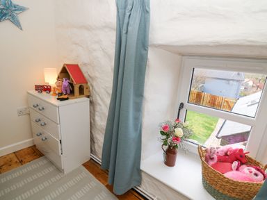 A corner of a room with a white chest of drawers with toys on top a window with blue curtains a vase with flowers and a basket with pink toys on the windowsill at Dyffryn Cottage in Aberporth