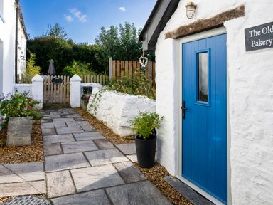 A paved path leading to a wooden gate with plants on both sides and a blue door on a white building at The Old Bakery