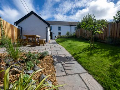 A garden with a stone path a wooden picnic table gravel area and a lawn with young trees at Dyffryn Cottage in Aberporth