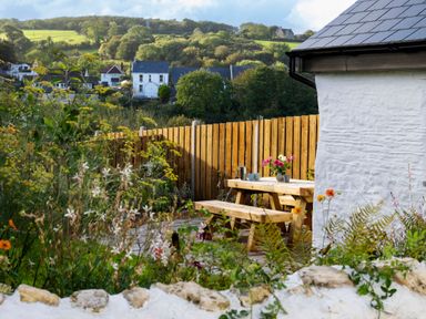 An outdoor wooden picnic table with benches near a white stone building and wooden fence with flowers and plants in the garden at Dyffryn Cottage in Aberporth