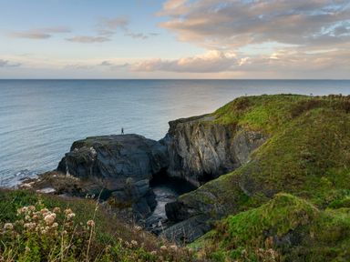 Rocky coastline with grassy hills and a person standing on a large rock by the sea at Dyffryn Cottage in Aberporth
