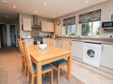 A kitchen with wooden dining table and chairs washing machine oven microwave and windows with patterned blinds at Beech Lodge in Goosnargh