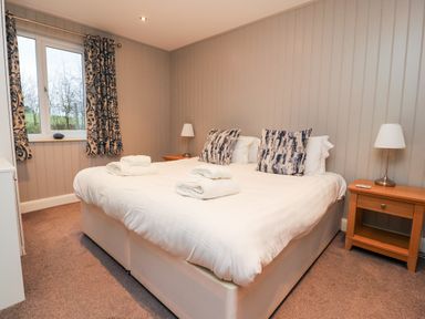 A bedroom with a double bed with two patterned pillows and folded towels two wooden bedside tables with lamps and a window with patterned curtains at Beech Lodge in Goosnargh