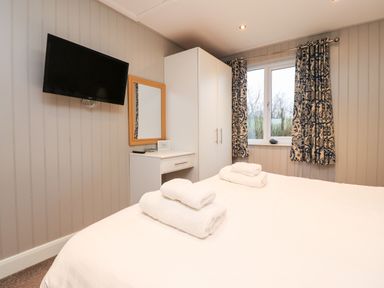 A bedroom with a bed, folded towels, a mounted TV, a wardrobe, a small dresser with a mirror, and a window with patterned curtains at Beech Lodge in Goosnargh