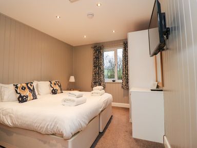 A bedroom with a double bed two folded towels bedside table with a lamp window with patterned curtains and a wall-mounted TV at Beech Lodge in Goosnargh
