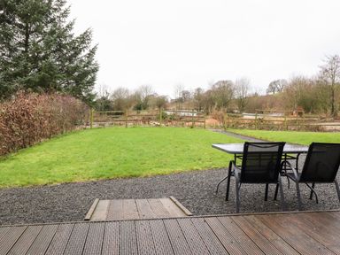 A backyard with green lawn a gravel area with table and two chairs and wooden deck at Beech Lodge in Goosnargh
