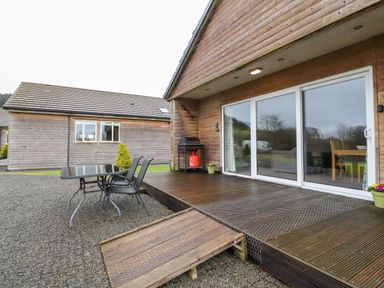 A wooden patio with a glass table and two chairs next to a building with sliding glass doors at Beech Lodge in Goosnargh