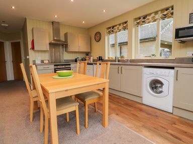A kitchen with a wooden dining table and four chairs a washing machine under the counter and three windows with patterned blinds at Oak Lodge in Goosnargh