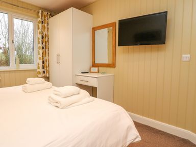 A bedroom with a bed with towels on it a wardrobe curtains a window a wall mirror and a wall mounted tv at Oak Lodge in Goosnargh