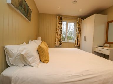 A bedroom with a large bed white pillows and yellow cushions a window with patterned curtains a white wardrobe and a dressing table with a mirror at Oak Lodge in Goosnargh