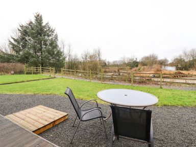 A patio area with a round table and two chairs overlooking a fenced grassy yard and trees at Oak Lodge in Goosnargh