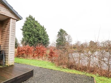A wooden building corner with stacked chairs on a patio near grass and shrubs with trees in the background at Oak Lodge in Goosnargh