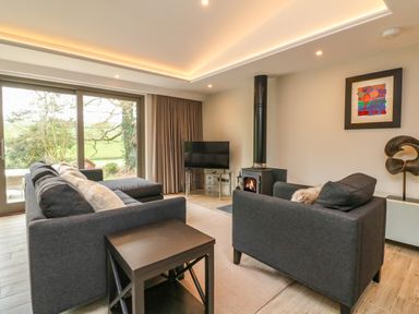 A living room with a dark grey sofa, armchair, wood stove, television, window with curtains and abstract wall art at Court Lodge in Bathealton near Wiveliscombe