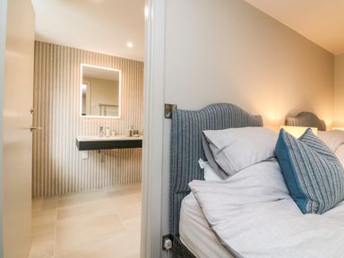 A bedroom with a blue upholstered headboard and pillows next to a bathroom with a lit mirror at Court Lodge in Bathealton near Wiveliscombe
