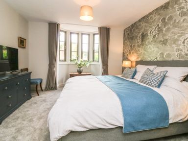 A bedroom with a large bed blue runner and pillows a chest of drawers and a window with curtains at Court Lodge Bathealton near Wiveliscombe