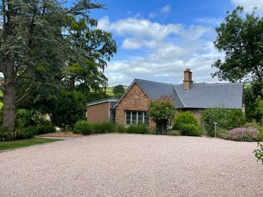 A stone house with a slate roof surrounded by trees and bushes with a gravel driveway at Court Lodge in Bathealton near Wiveliscombe