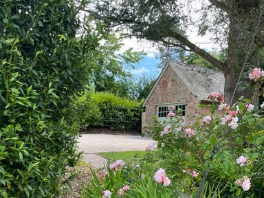 A stone building with a pitched roof surrounded by green bushes and pink flowering plants at Court Lodge in Bathealton near Wiveliscombe