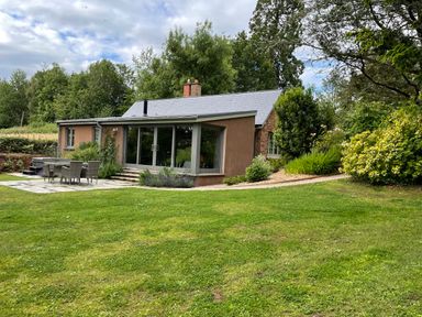 A house with large windows and patio furniture on a lawn surrounded by trees and bushes at Court Lodge in Bathealton near Wiveliscombe