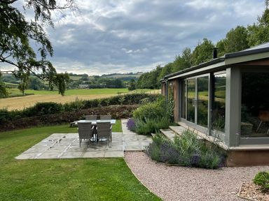A patio with a dining table and chairs next to a house with large windows and a garden at Court Lodge in Bathealton near Wiveliscombe