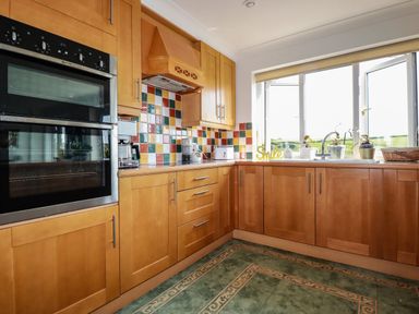 A kitchen with wooden cabinets, built-in oven, colorful tiled backsplash, sink under a window, and small appliances on the countertop at Newquay Tamarisk Lodge on the Gannel in Newquay