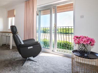 A room with a black reclining chair a small table with pink flowers and glass sliding doors opening to a balcony overlooking greenery and water at Newquay Tamarisk Lodge on the Gannel in Newquay