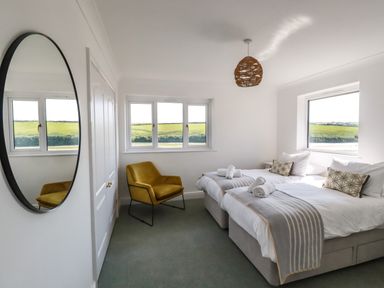 A bedroom with two single beds a yellow chair a round mirror and windows showing green fields at Newquay Tamarisk Lodge on the Gannel in Newquay
