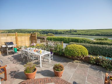 An outdoor patio with a dining table and chairs set with food and drinks a barbecue grill potted plants and a view of greenery and a river at Newquay Tamarisk Lodge on the Gannel in Newquay