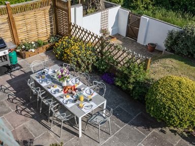 An outdoor patio with a dining table set for breakfast surrounded by chairs and garden plants at Newquay Tamarisk Lodge on the Gannel Newquay