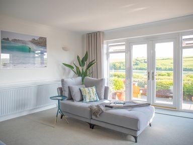 A room with a gray chaise lounge next to glass doors showing greenery outside at Newquay Tamarisk Lodge on the Gannel in Newquay