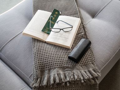 A grey throw with fringes on a grey couch with an open book a green bookmark a pair of glasses and a glasses case on top at Newquay Tamarisk Lodge on the Gannel in Newquay