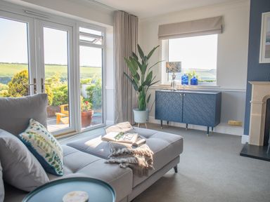 A living room with a gray couch a blue cabinet a potted plant and glass doors leading outside at Newquay Tamarisk Lodge on the Gannel in Newquay
