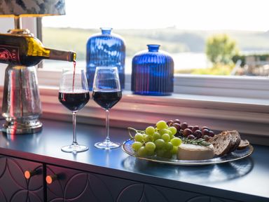 A counter with two glasses of red wine being poured a plate with grapes cheese and bread and two blue glass vases at Newquay Tamarisk Lodge on the Gannel in Newquay