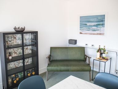 A sitting area with a green bench a black cabinet with glass doors and a small side table with wine and a photo of the ocean on the wall at Newquay Tamarisk Lodge on the Gannel in Newquay