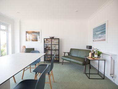 A dining area with a white table surrounded by blue chairs a green sofa a glass cabinet and wall art at Newquay Tamarisk Lodge on the Gannel in Newquay