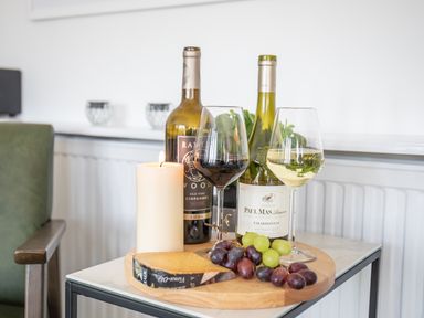 A wooden tray with two wine bottles two wine glasses grapes cheese and a lit candle on a side table in a room at Newquay Tamarisk Lodge on the Gannel in Newquay