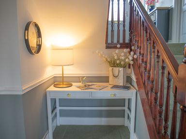 A hallway corner with a white table holding a lamp, books, and a flower vase next to a wooden staircase at Newquay Tamarisk Lodge on the Gannel in Newquay